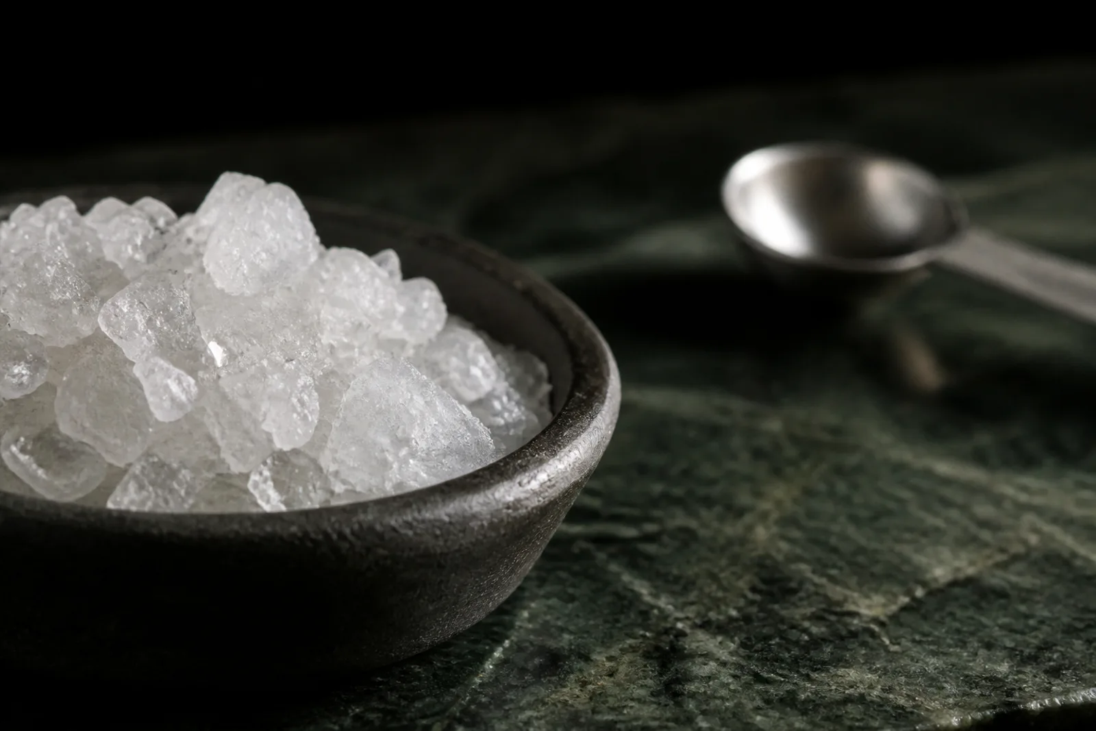 Coarse white aquarium salt crystals in a plain dish beside a measuring spoon on a neutral surface